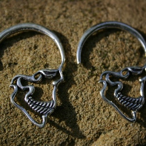 May include: A pair of silver skull-shaped earrings with intricate details. The earrings are made of metal and have a circular shape.
