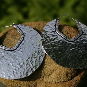 May include: A pair of large, silver hoop earrings with a hammered texture. The earrings are round and have a slightly curved shape.