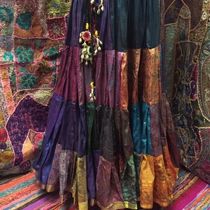 May include: A long, tiered skirt in a patchwork design, featuring a variety of colors including purple, teal, orange, and gold. The skirt has a black braided belt with decorative beaded tassels. The skirt appears to be made of silk or a similar fabric.