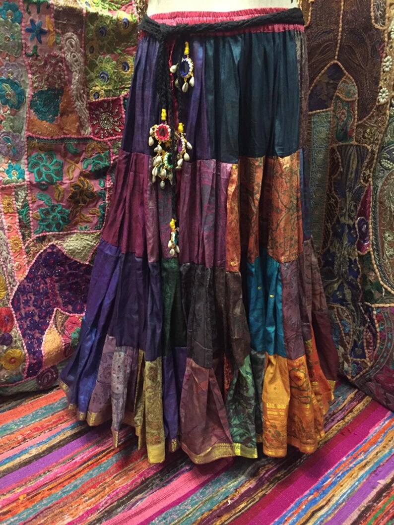 May include: A long, tiered skirt in a patchwork design, featuring a variety of colors including purple, teal, orange, and gold. The skirt has a black braided belt with decorative beaded tassels. The skirt appears to be made of silk or a similar fabric.