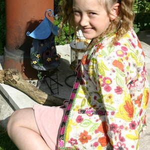 May include: A young girl with pigtails wears a white floral jacket with a pink skirt. She is sitting on a concrete surface with a small blue and white metal fairy house in the background.