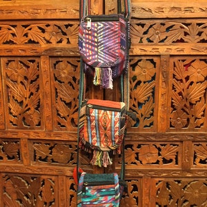 Pode incluir: Tr&ecirc;s pequenas bolsas coloridas e estampadas com franjas penduradas na parte inferior. As bolsas est&atilde;o presas a al&ccedil;as longas e est&atilde;o penduradas em uma parede de madeira.
