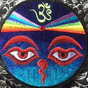 Peut inclure: Un patch rond brodé avec un fond bleu et un arc-en-ciel en haut. Le patch présente un symbole Om vert, deux yeux rouges avec des pupilles blanches et une forme spirale rouge au centre.