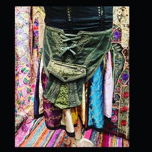 May include: A green canvas belt with a lace-up front and a zippered pocket. The belt is decorated with embroidered details and is worn over a colorful skirt.