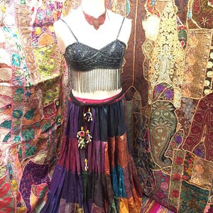 May include: A mannequin displays a bohemian-style outfit. The ensemble includes a beaded black crop top with fringe, a tiered patchwork skirt in various colors, and a matching necklace. The background features colorful textiles.