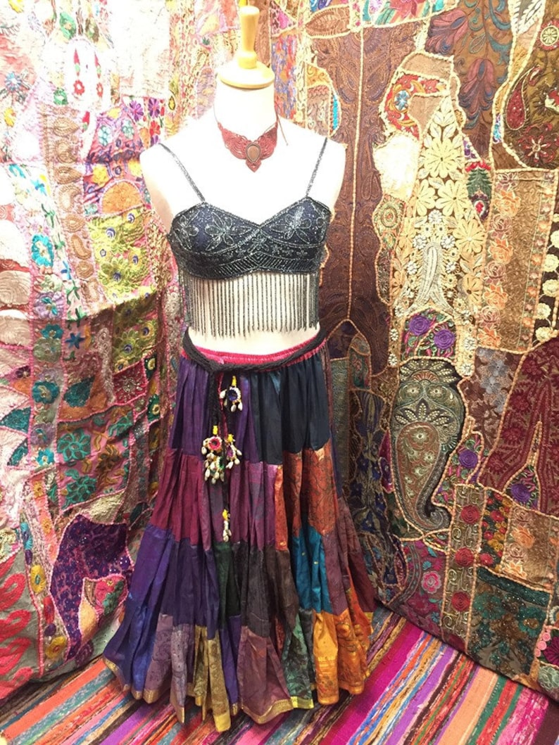 May include: A mannequin displays a bohemian-style outfit. The ensemble includes a beaded black crop top with fringe, a tiered patchwork skirt in various colors, and a matching necklace. The background features colorful textiles.