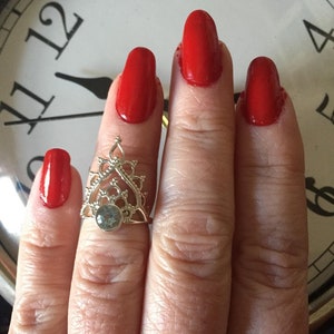 May include: A silver ring with a delicate design and a blue gemstone. The ring is worn on the middle finger of a hand with red nail polish.