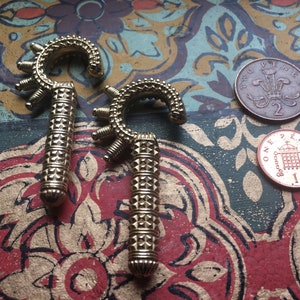 May include: Two gold-colored metal hooks with intricate designs. The hooks have a rounded top and a pointed bottom. The hooks are on a patterned surface.