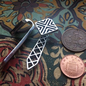 May include: Two silver tie clips with geometric designs, one with a square shape and the other with a long, narrow shape. The tie clips are lying on a colorful patterned surface with two British coins, one penny and two pence.