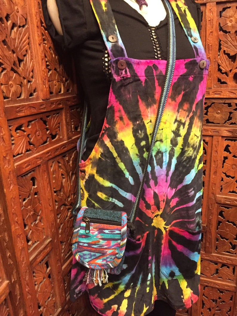 Pode incluir: Um vestido de macac&atilde;o tie-dye com uma pequena bolsa transversal tecida colorida. O vestido tem um fundo preto com redemoinhos tie-dye arco-&iacute;ris.