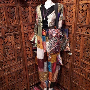 May include: A patchwork pantsuit with a long, flowing jacket and a matching tote bag. The fabric is a mix of colourful prints and textures, including paisley, floral, and geometric patterns. The outfit is accessorised with a silver necklace and a black belt.