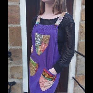 Könnte beinhalten: Lila Cord-Latzkleid mit Patchwork-Taschen und Trägern. Das Kleid hat eine lockere Passform und dekorative Patches in verschiedenen Farben und Mustern. Die Träger sind mit Knöpfen befestigt. Die Website-Adresse www.gutsygingers.etsy.com befindet sich unten.