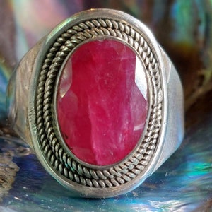 Puede incluir: Un anillo de plata con una gran piedra preciosa rosa de forma ovalada engastada en un engaste de plata. El anillo tiene un borde decorativo trenzado alrededor de la piedra preciosa.