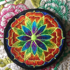 Peut inclure: Un patch rond brodé avec un motif de mandala coloré. Le mandala présente un arc-en-ciel de couleurs, notamment le rouge, l'orange, le jaune, le vert, le bleu, l'indigo et le violet. Le centre du mandala est un cercle vert vif.