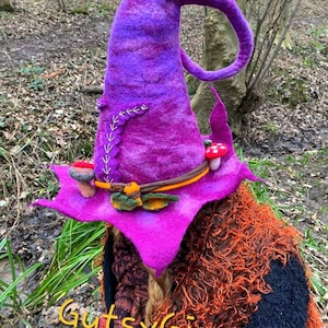 Peut inclure: Un chapeau de sorcière violet vif avec une pointe recourbée, orné d'une bande décorative et de petits champignons. Le chapeau présente un bord pointu et des détails cousus. Le texte "GutsyGingers" est visible en bas.