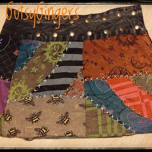 Puede incluir: Una falda de retazos con una cintura de mezclilla negra y tachuelas plateadas. La falda está hecha de diferentes telas con varios patrones, incluyendo abejas, estrellas y lunas. La falda es una prenda única y elegante.