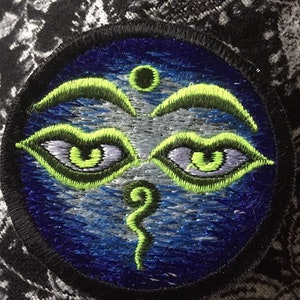 May include: A round, blue and green embroidered patch with a third eye design. The eye is surrounded by a green eyebrow and a green swirl.