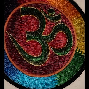 May include: A circular embroidered patch with a green Om symbol in the center. The Om symbol is surrounded by a rainbow of colors, including red, orange, yellow, green, blue, and purple. The patch is edged in black.