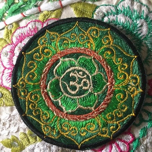Peut inclure: Écusson brodé circulaire avec un motif détaillé en vert, or et rouge. Le patch a une bordure noire et présente le symbole Om au centre. Il est posé sur un tissu brodé blanc et floral.