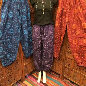 Può includere: Tre paia di pantaloni stampati con un designo di sole e luna. I pantaloni sono di colore blu, viola e arancione. I pantaloni sono appesi a un rack di legno.