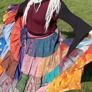 May include: A woman wearing a burgundy top with long black sleeves and a colorful patchwork skirt. The skirt features various fabrics in shades of blue, orange, pink, and purple. The woman has long, white dreadlocks and is outdoors on a grassy area.