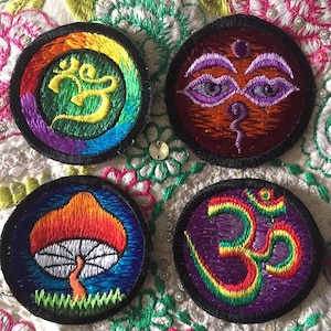 Peut inclure: Quatre patchs brodés avec des motifs différents. Le premier patch est un cercle arc-en-ciel avec un symbole Om au centre. Le deuxième patch est un cercle brun avec un motif de troisième œil violet. Le troisième patch est un cercle bleu avec un motif de champignon rouge et orange. Le quatrième patch est un cercle violet avec un symbole Om jaune et rouge au centre.