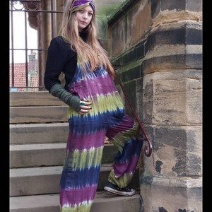 May include: A woman wearing a black mock neck shirt and tie-dye overalls with a purple, green, and blue color scheme. She is wearing a purple headband and black and white sneakers. The woman is standing on a set of stone steps with a stone wall behind her.