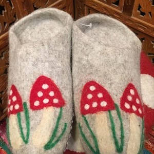 May include: Grey felt slippers decorated with a mushroom motif. Each slipper has three red mushrooms with white spots and green stems. The slippers are made from a soft, textured material.