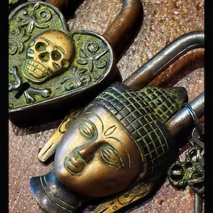 May include: Two antique brass padlocks with intricate designs. One padlock is heart-shaped with a skull and crossbones, the other is a Buddha head.