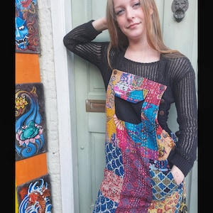 May include: A woman wearing a black jumper over a colourful patchwork overall dress. The dress has a large pocket on the front and is made of different fabrics. The dress is a unique and stylish piece of clothing.