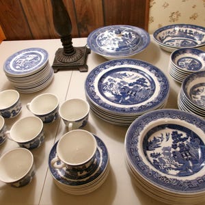 Op de afbeelding: Een grote set blauw-wit keramisch servies met een gedetailleerd wilgenpatroon. De set bevat stapels dinerborden, saladeborden, kommen en kopjes met bijpassende schoteltjes, allemaal versierd met ingewikkelde landschapstaferelen.