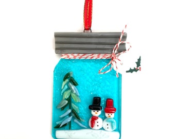 May include: A glass ornament shaped like a mason jar with a blue background, a white snowy base, a green Christmas tree, and two snowmen with black hats and red scarves.