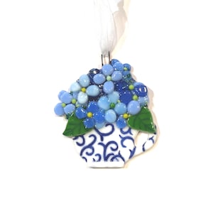 May include: A handcrafted glass ornament shaped like a teacup filled with blue hydrangea flowers. The teacup is white with blue scrollwork. The flowers are various shades of blue with green leaves. A white ribbon is attached for hanging.