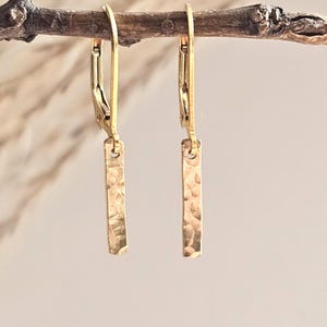 May include: A pair of gold-toned drop earrings featuring a vertical rectangular bar pendant with a hammered, textured finish. Each earring is attached to a hinged lever-back closure, creating a simple and clean design with a metallic luster.