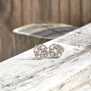May include: A pair of silver stud earrings featuring a circular Celtic knot design. Each earring is crafted from polished metal with an intricate, interwoven pattern that forms a continuous, rounded shape, showcasing a classic, symmetrical knotwork style.