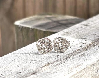 Puede incluir: Un par de pendientes de botón de plata con un diseño de nudo celta circular. Cada pendiente está elaborado en metal pulido con un patrón intrincado y entrelazado que forma una figura redondeada continua, mostrando un estilo simétrico.