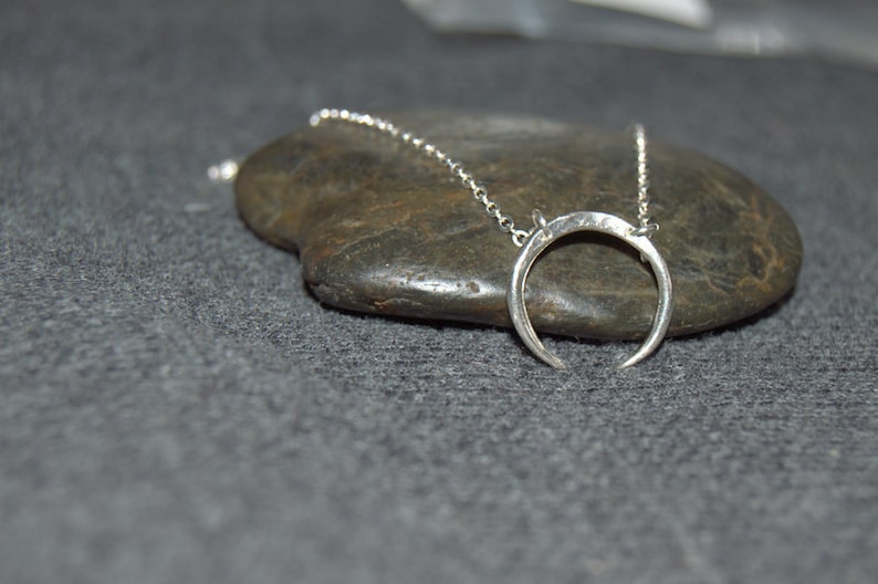 May include: A silver crescent moon pendant necklace on a silver chain, resting on a gray rock.