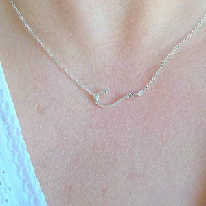May include: A silver-toned necklace featuring a delicate chain and a central fish hook pendant. The metal hook is shaped with a curved point and a small barb, connected directly to the thin chain links on both sides, creating a simple, continuous design.