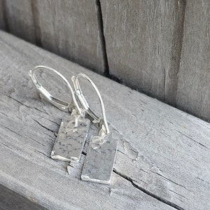 May include: A pair of silver earrings with hammered rectangular pendants hanging from silver hoops.
