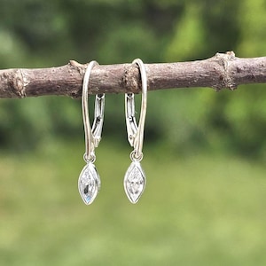 May include: A pair of silver-toned leverback earrings, each featuring a single marquise-cut clear gemstone set in a bezel frame. The earrings hang from a thin, curved metal wire, showcasing a simple and refined design with polished metal.