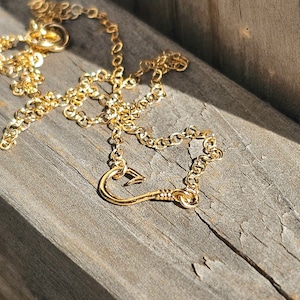 May include: A gold chain necklace with a heart-shaped hook pendant. The necklace is lying on a wooden surface.