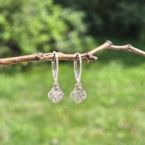 May include: A pair of silver-toned drop earrings featuring a small, intricate, four-lobed Celtic knot design. The earrings are suspended from simple lever-back closures, showcasing a polished metallic finish and a symmetrical, woven pattern.