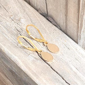 May include: A pair of gold-toned leverback earrings featuring small, smooth, oval-shaped pendants. The earrings have a polished metallic finish and a simple, clean design with a secure hinged closure mechanism for each earring piece.
