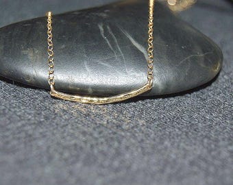 May include: A gold-toned necklace featuring a delicate chain and a curved, textured bar pendant. The metal surface of the bar has a hammered, organic finish, and the chain links are small and circular, creating a simple and refined jewelry piece.