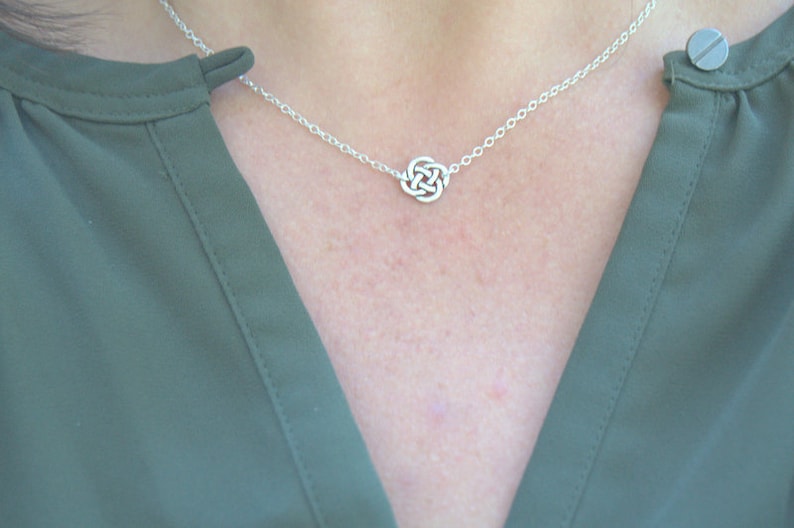 May include: A silver chain necklace with a small, intricate Celtic knot pendant. The necklace is worn by a person wearing a green shirt.