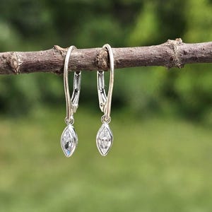 May include: A pair of silver-toned leverback earrings featuring a marquise-cut clear stone set in a bezel setting. The earrings hang from a thin, curved metal wire, showcasing a polished metallic finish and a faceted, elongated gemstone design.