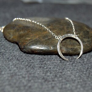 May include: A silver crescent moon pendant necklace on a silver chain. The necklace is displayed on a gray rock.