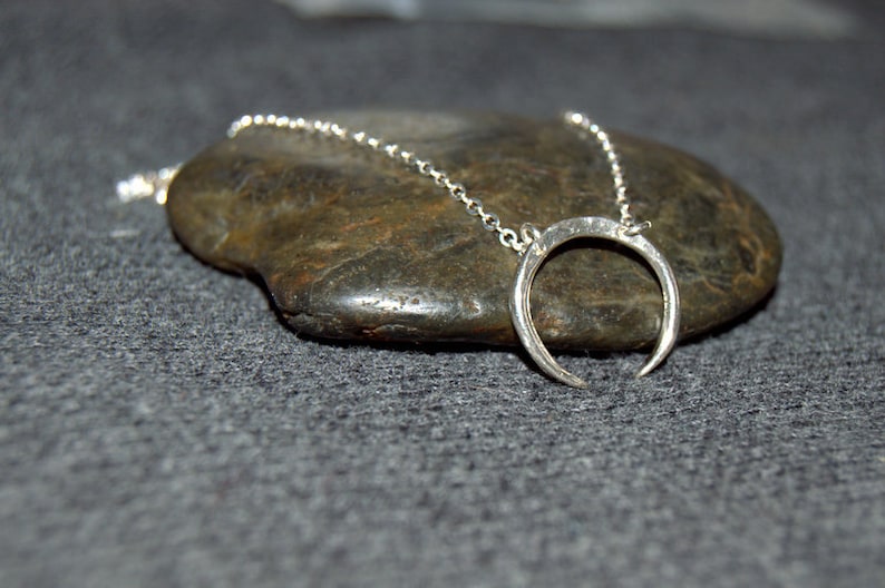 May include: A silver crescent moon pendant necklace on a silver chain. The necklace is displayed on a gray rock.