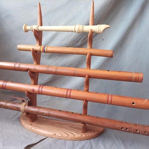 May include: A wooden display stand holding five flutes. The flutes vary in color and design, with some featuring decorative elements. The stand is made of light brown wood and has a rounded base.