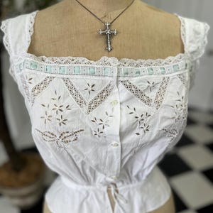 Puede incluir: Una blusa de algod&oacute;n blanco con delicado ribete de encaje y detalles florales bordados. La blusa tiene un frente abotonado y una cintura ajustada.